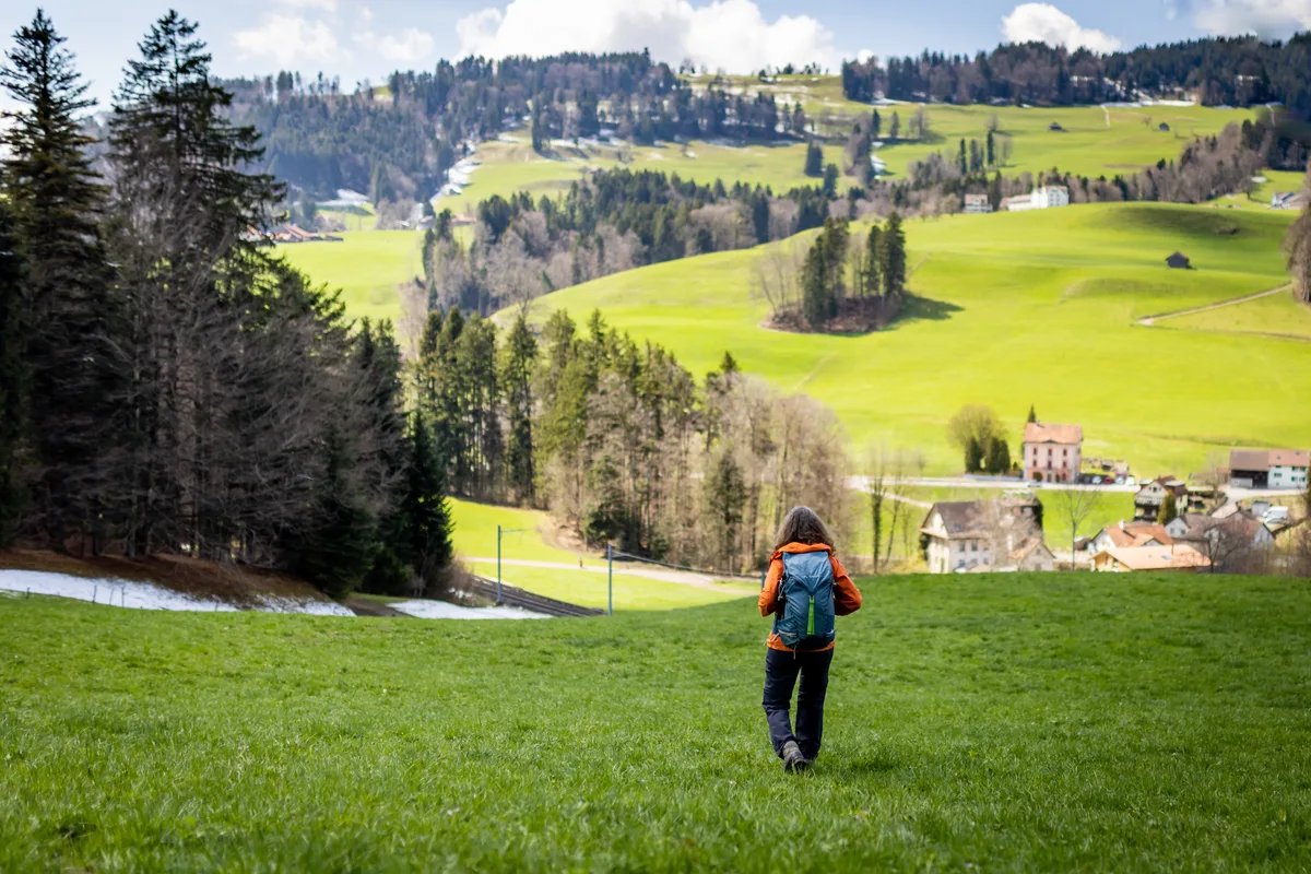 Eine Frau in oranger Jacke und mit blauem Rucksack wandert über eine grüne Wiese.